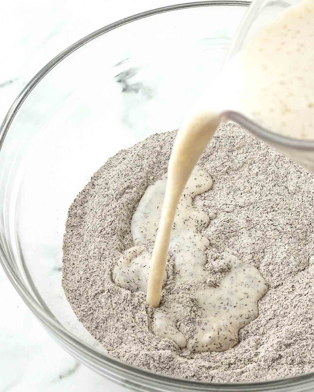 Wet ingredients being poured into a glass bowl of dry ingredients.