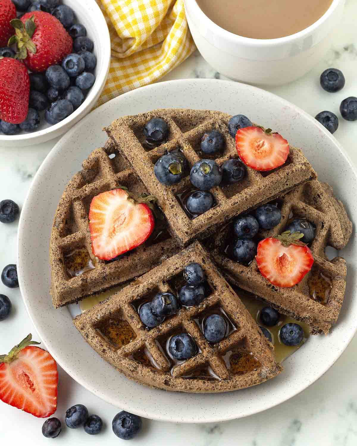An overhead image showing a buckwheat waffle on a plate, the waffle has syrup and berries on top of it.