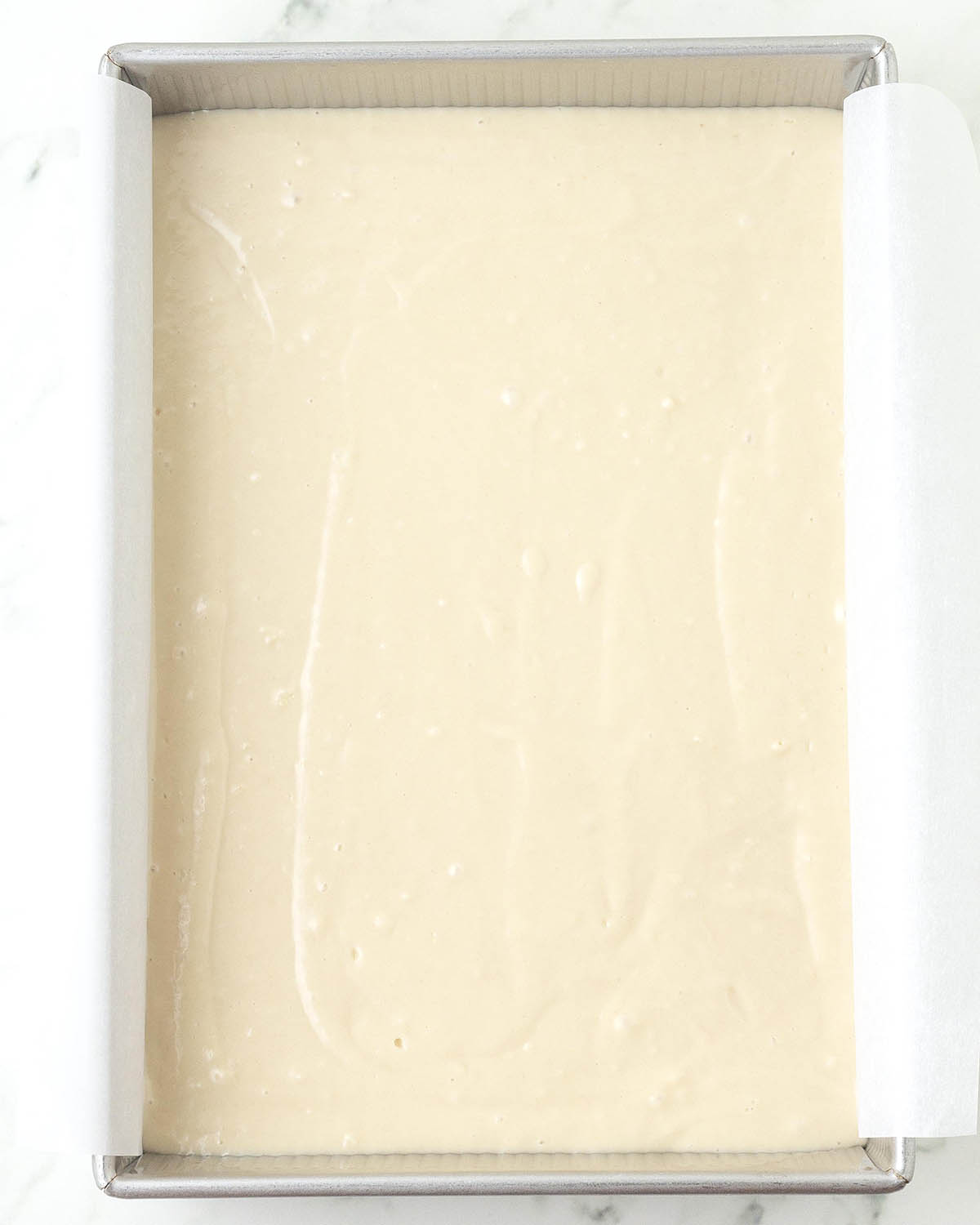 Unbaked vanilla sheet cake batter in a rectangle, parchment lined baking pan.