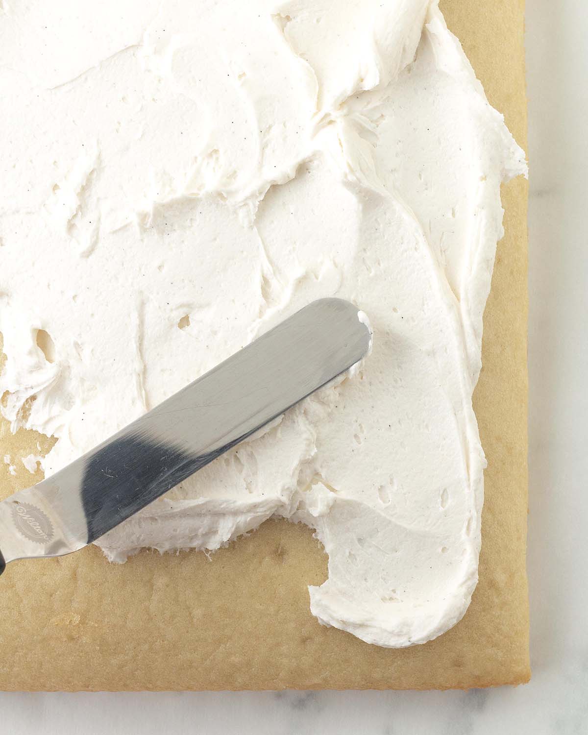 A small offset spatula spreading vanilla frosting onto a vanilla sheet cake.