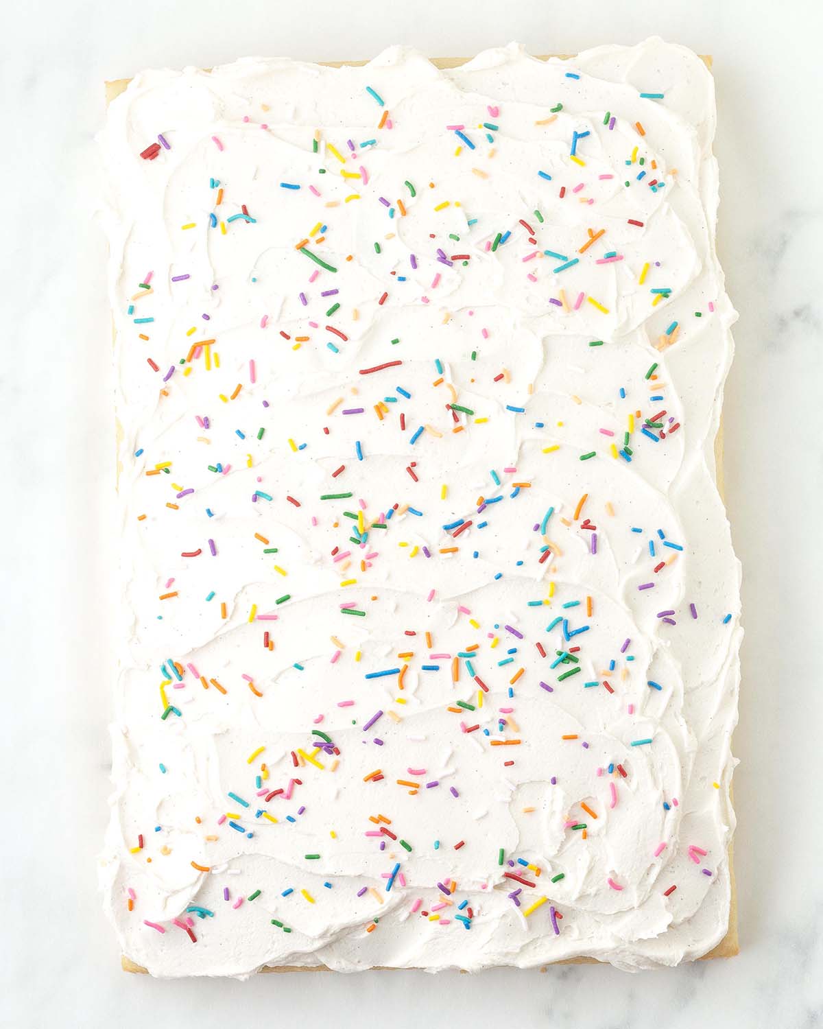 An overhead image showing a vanilla sheet cake that has been topped with vanilla frosting and rainbow sprinkles.