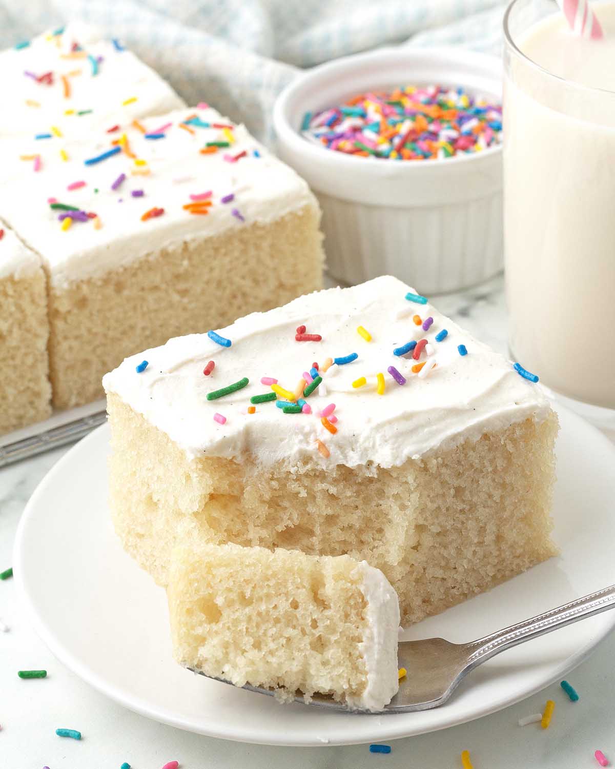A piece of vegan vanilla sheet cake on a white plate, a fork with a piece of the cake on it sits in front of the cake.