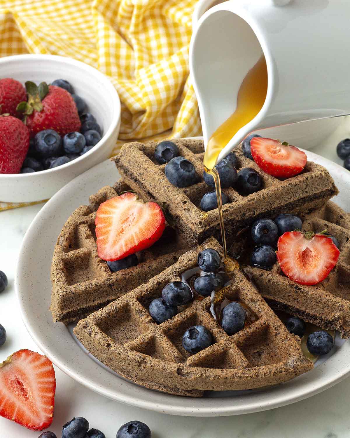 Maple syrup being poured onto a waffle that is topped with fresh blueberries and strawberries.